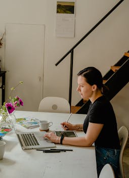 Woman using a digital pen with her laptop in a creative workspace featuring art materials.